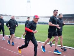 A few men jog on a track, smailing and mostly wearing black