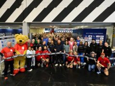 A large group of people and a huge teddy bear dressed in running kit pose in front of the ribbon to formally open the show