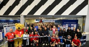 A large group of people and a huge teddy bear dressed in running kit pose in front of the ribbon to formally open the show