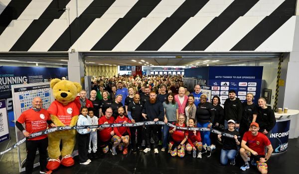 A large group of people and a huge teddy bear dressed in running kit pose in front of the ribbon to formally open the show