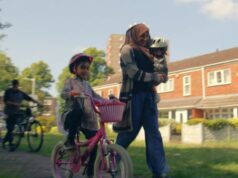 A lady with a head covering running with her family of young children, one is a babe in her arms, two are on bikes, and a man on a bike is behind with one of the children