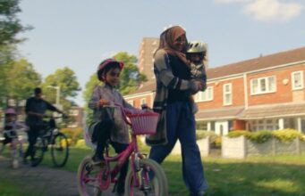 A lady with a head covering running with her family of young children, one is a babe in her arms, two are on bikes, and a man on a bike is behind with one of the children