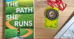 Photo shows copy of the Path She Runs on a wooden table next to a map and compass
