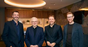 4 men all wearing black or dark suits stand in a room and smile for the camera