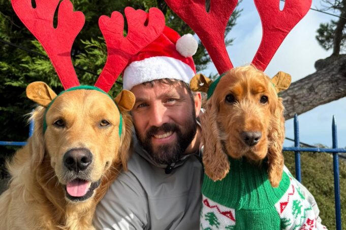 Man wearing santa hat poses wiith two golden dogs dressed as reindeer