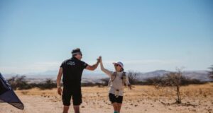 Man in black shorts and T-shirt hi-fives a woman in the desert.
