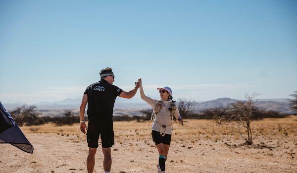 Man in black shorts and T-shirt hi-fives a woman in the desert.
