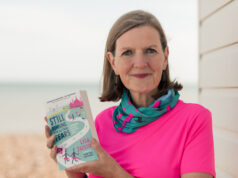 Lady with short brown hair in a pink t shirt and green and pink neck warer holds a book
