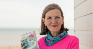 Lady with short brown hair in a pink t shirt and green and pink neck warer holds a book
