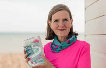 Lady with short brown hair in a pink t shirt and green and pink neck warer holds a book