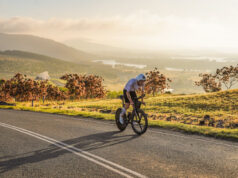 Man cycles on a road overlooking a beautiful green valley with a lake in the distance