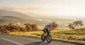 Man cycles on a road overlooking a beautiful green valley with a lake in the distance