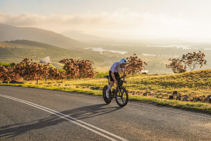 Man cycles on a road overlooking a beautiful green valley with a lake in the distance