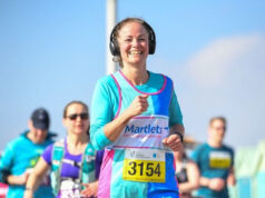 Woman in blue top and vest wearing big black headphones smiles as she runs. The sky is blue, and other runners are also visible.