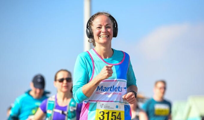 Charity places available for Brighton Half Woman in blue top and vest wearing big black headphones smiles as she runs. The sky is blue, and other runners are also visible.