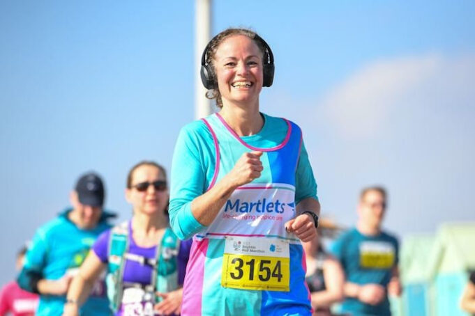 Woman in blue top and vest wearing big black headphones smiles as she runs. The sky is blue, and other runners are also visible.