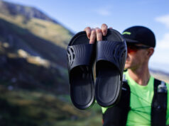 Male runner wearing black cap, green t-shirt and black race vest holds up a pair of black recovery sliders.