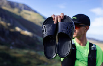 Enertor challenges London’s running community Male runner wearing black cap, green t-shirt and black race vest holds up a pair of black recovery sliders.