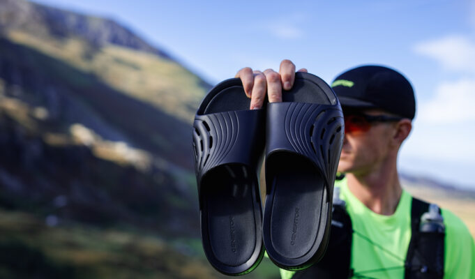 Enertor challenges London’s running community Male runner wearing black cap, green t-shirt and black race vest holds up a pair of black recovery sliders.