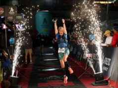IRONMAN 2026 schedule for US and Canada Woman jumps for joy across and Ironman finish line. It's night time and there are sparkling lights showering beside her.
