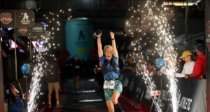 Woman jumps for joy across and Ironman finish line. It's night time and there are sparkling lights showering beside her.