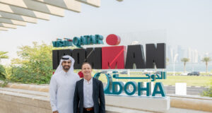 Two men stand in front of IRONMAN Doha sign. Man on right is dressed in black suit, man on left is wearing a white thawb