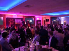 Phooto of a room full of people dressed smartly, at round tables, all watching a speaker on a stage