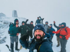 Numerous men cheer with arms in the air atop a snowy mountain with a trig pillar in the background.