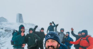 Numerous men cheer with arms in the air atop a snowy mountain with a trig pillar in the background.