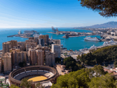 A cityscape by a coastline. Tall buildings and a bull ring are visible, as it a wooded hillside and blue sea