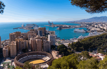 A cityscape by a coastline. Tall buildings and a bull ring are visible, as it a wooded hillside and blue sea