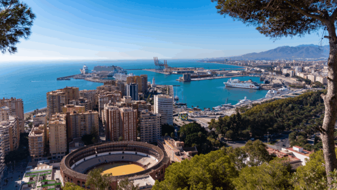 A cityscape by a coastline. Tall buildings and a bull ring are visible, as it a wooded hillside and blue sea