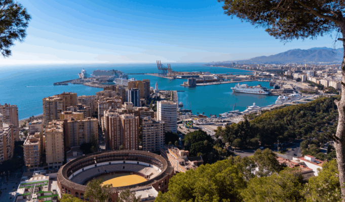 A cityscape by a coastline. Tall buildings and a bull ring are visible, as it a wooded hillside and blue sea