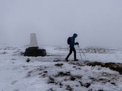 A lone walker in winter kit passes a lone trig point in a snowy, foggy landscape