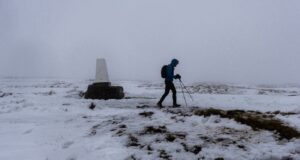 A lone walker in winter kit passes a lone trig point in a snowy, foggy landscape