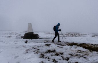 A lone walker in winter kit passes a lone trig point in a snowy, foggy landscape