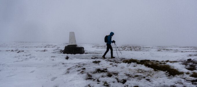 Credit: David Murch / The Spine Race A lone walker in winter kit passes a lone trig point in a snowy, foggy landscape
