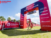 A single triathlete walks under a finish gantry with the finish tape above their head.