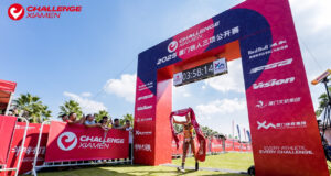 A single triathlete walks under a finish gantry with the finish tape above their head.