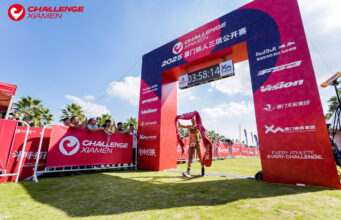 A single triathlete walks under a finish gantry with the finish tape above their head.