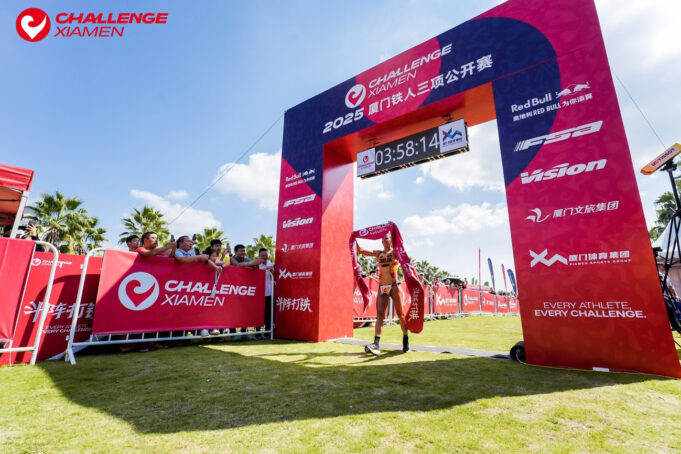 A single triathlete walks under a finish gantry with the finish tape above their head.