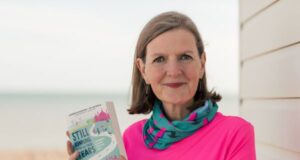 A woman with brown shoulder length hair wearing a pink t-shirt and a neck warmer with flamingos on it holds her book Still Running After All These Tears