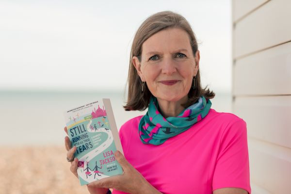 A woman with brown shoulder length hair wearing a pink t-shirt and a neck warmer with flamingos on it holds her book Still Running After All These Tears