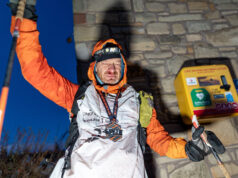Sebastien Raichon wins Montane Winter Spine A man stands at the wall of a building next to a defibrillator. He is wearing full winter kit and wearing a medal, and holds one of his poles in the air. He looks physically exhausted.