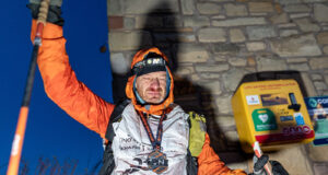 A man stands at the wall of a building next to a defibrillator. He is wearing full winter kit and wearing a medal, and holds one of his poles in the air. He looks physically exhausted.