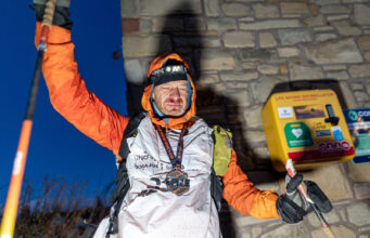 A man stands at the wall of a building next to a defibrillator. He is wearing full winter kit and wearing a medal, and holds one of his poles in the air. He looks physically exhausted.