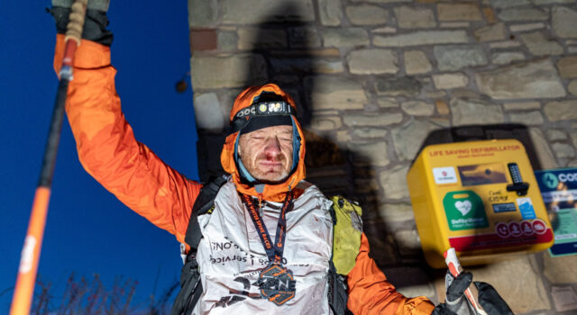 Sebastien Raichon wins Montane Winter Spine A man stands at the wall of a building next to a defibrillator. He is wearing full winter kit and wearing a medal, and holds one of his poles in the air. He looks physically exhausted.
