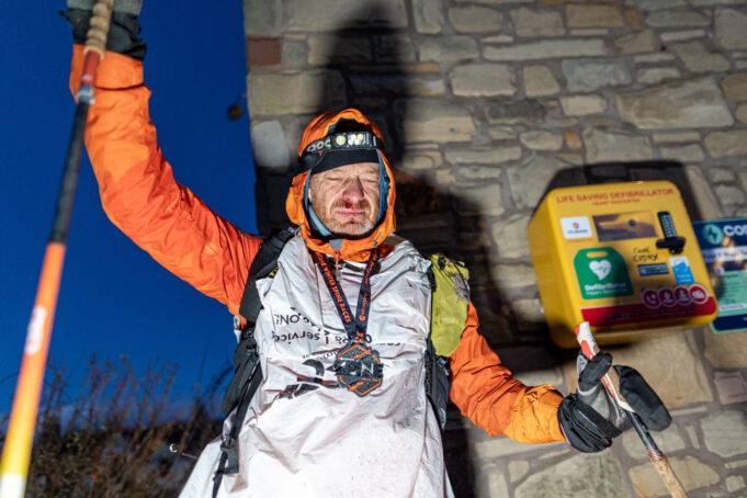 A man stands at the wall of a building next to a defibrillator. He is wearing full winter kit and wearing a medal, and holds one of his poles in the air. He looks physically exhausted.