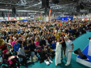 A huge crowd of people watch a stage with Harry Judd, Susie Chan and Ewan Thomas, who are all taking a selfie of themselves with the crowd
