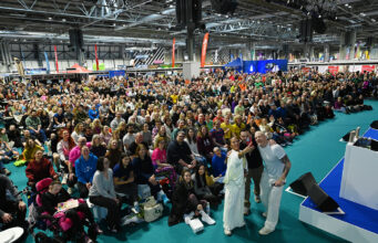 A huge crowd of people watch a stage with Harry Judd, Susie Chan and Ewan Thomas, who are all taking a selfie of themselves with the crowd